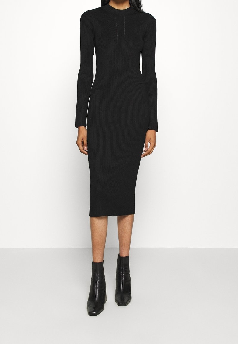 Schwarzes geripptes Kleid mit langen Ärmeln und hochgeschlossenem Kragen, das eine figurbetonte Silhouette und einen knielangen Saum hat. Kombiniert mit schwarzen Knöchelstiefeln.