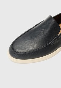 Chaussure noire en cuir sans lacets avec semelle en caoutchouc blanche, bout arrondi et détail de grain texturé sur la tige.