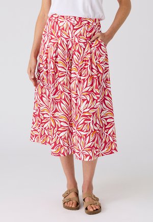 Frau trägt einen roten Midirock mit weißen und gelben Blattmustern, kombiniert mit beigen Sandalen mit Schnalle und einem weißen Oberteil.