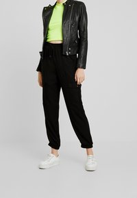 Svart lädermotorcykeljacka, neongrön kort topp, svarta cargobyxor med fickor och vita sneakers med strukturerade gummisulor.