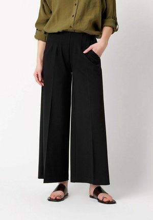 Person trägt lockere schwarze Hose mit vorderen Falten, olivgrünes hochgekrempeltes Hemd und schwarze offene Sandalen, steht mit einer Hand in der Tasche.