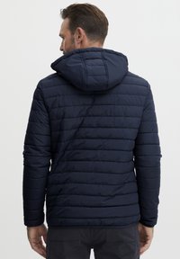 Jachetă pufoasă de marine, cu modele de cusătură orizontală, glugă și mansete elastice. Materialul ușor oferă un aspect elegant și ajustat.