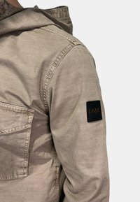 Giacca beige chiaro con una texture liscia, dotata di cappuccio, tasca sul petto e un patch del logo nero sulla manica.