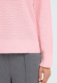 Hellrosa strukturierter Pullover mit geripptem Saum, der ein geschichtetes Strickdesign aufweist. Kombiniert mit grauen Hosen, wobei Hand- und Ärmeldetails sichtbar sind.