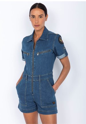 Salopette en denim bleu foncé avec des manches courtes, fermeture à glissière sur le devant, deux zips sur la poitrine et poches latérales. Comprend un badge sur la manche gauche.