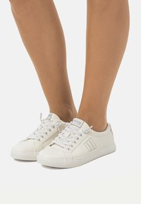 Zapatillas de lona blancas con diseño de cordones, punta redonda y un sutil detalle del logo en el costado, descansando sobre piernas desnudas. Textura suave.