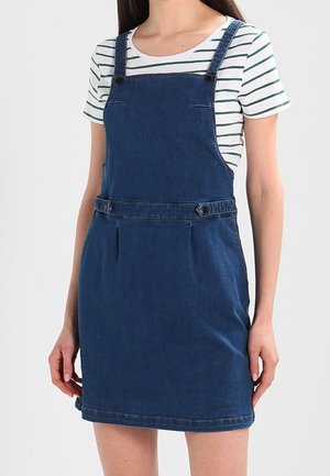 Kobieta w niebieskiej dżinsowej sukience na szelkach, nałożonej na białą bluzkę z krótkim rękawem i zielonymi poziomymi paskami, stoi na jednolitym tle.