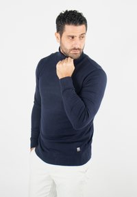 Pull à col roulé bleu marine en tissu tricoté, avec des poignets et un ourlet côtelés, présentant un petit logo sur le bas à gauche.