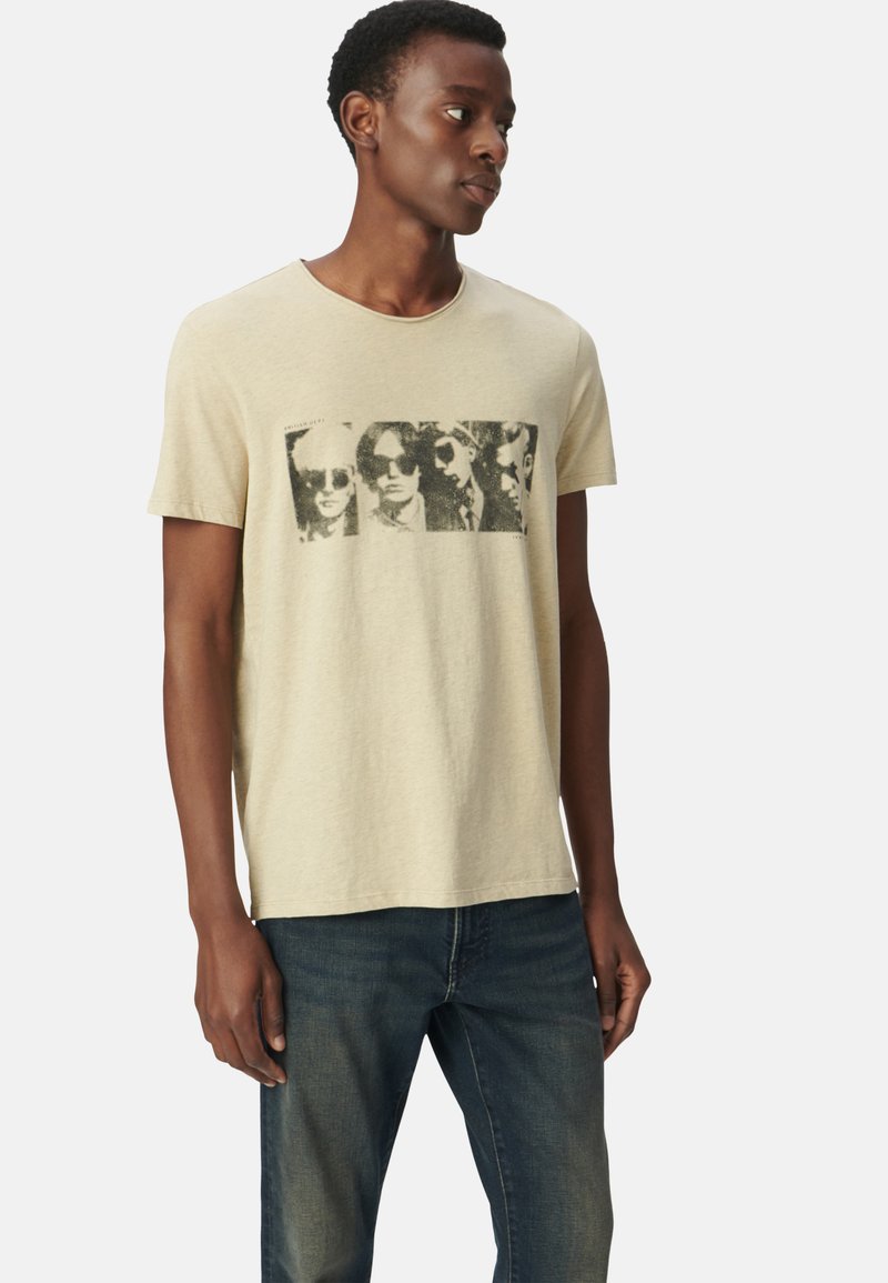 Camiseta de algodón beige con mangas cortas que presenta un gráfico en blanco y negro de cuatro rostros con gafas de sol. Cuello redondo y corte relajado.