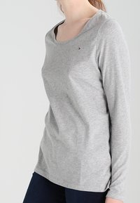 Grauer Langarm-T-Shirt mit Rundhalsausschnitt, aus weichem Material gefertigt, mit einem kleinen Logo-Akzent auf der Brust und einer lockeren Passform.