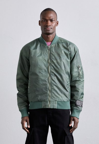 Jachetă bomber verde cu fermoar frontal, manșete și tiv ribbed, buzunare laterale și un patch decorativ pe mânecă. Finisaj neted, texturat.