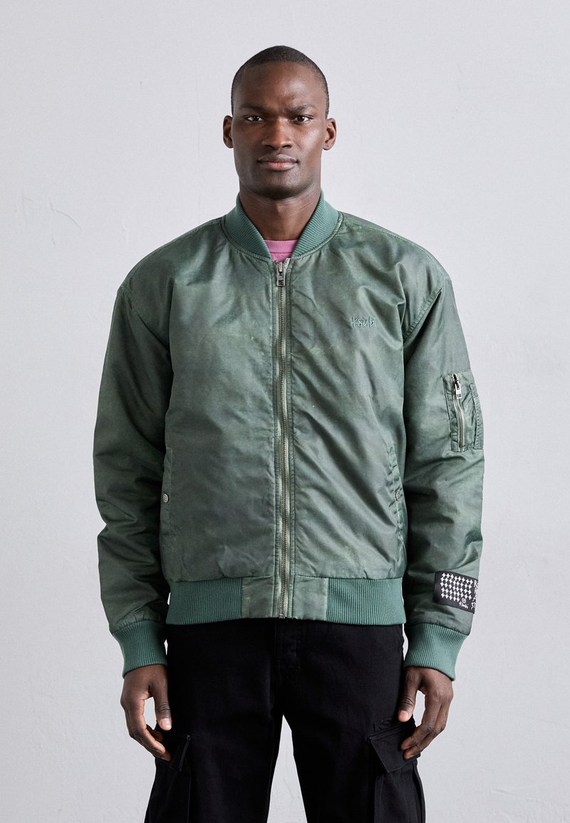 Veste bomber verte avec fermeture éclair à l'avant, poignets et ourlet côtelés, poches latérales et un patch décoratif sur la manche. Fini lisse ettexturé.