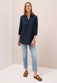 Tunique bleu marine à manches trois-quarts avec col, patte de boutonnage et fentes sur les côtés ; associée à un jean bleu clair et des sabots marron.
