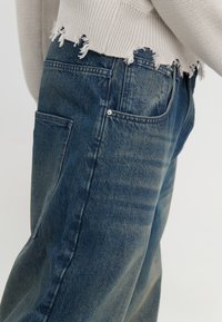 Vaqueros de mezclilla azul oscuro con textura desgastada, que cuentan con un bolsillo lateral y detalle de dobladillo deshilachado. El material parece resistente y casual.