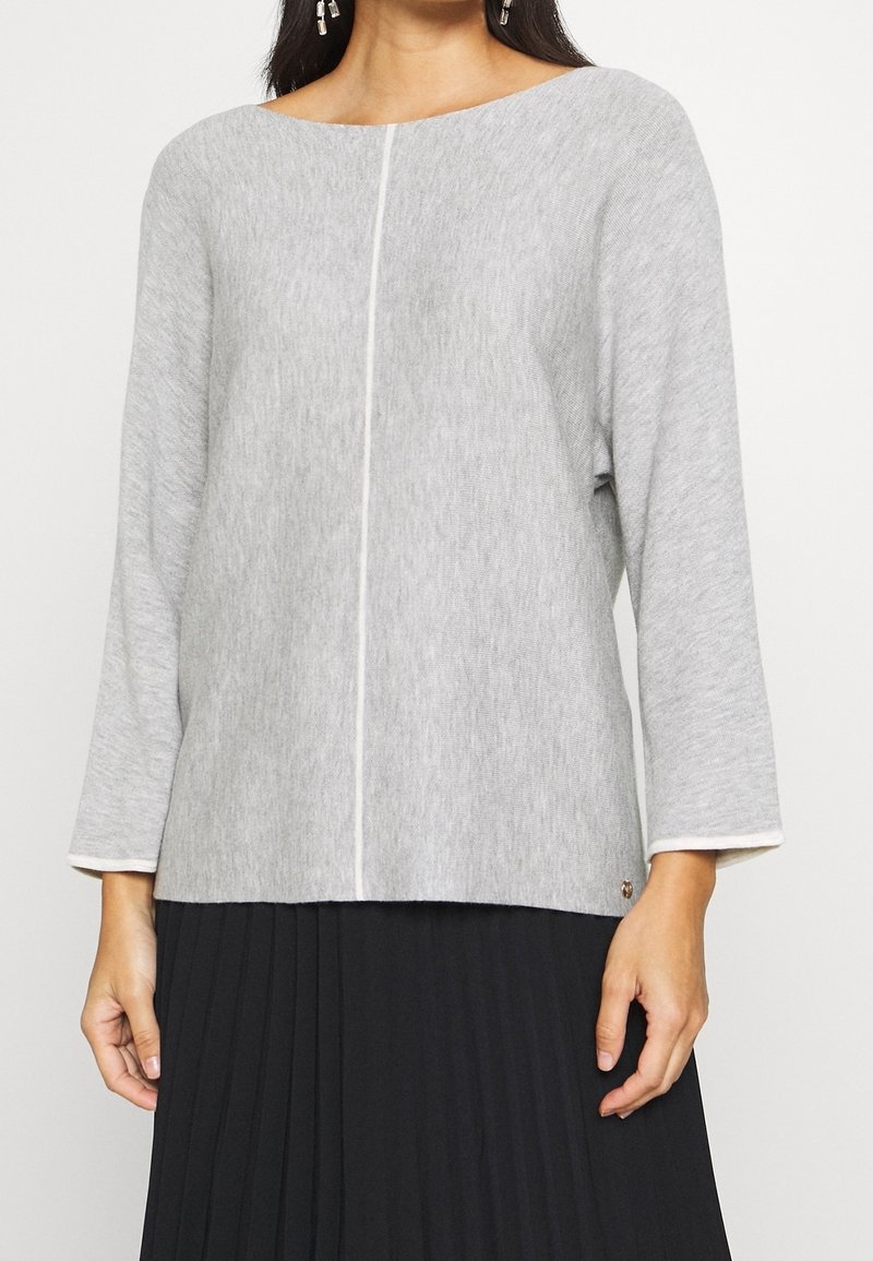 Femme portant un pull en maille gris clair à manches longues avec une couture centrale, associé à une jupe plissée noire.