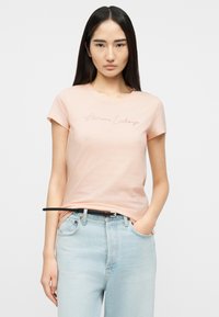 Νέα γυναίκα με μακριά σκούρα μαλλιά που φοράει ένα ροζ ανοιχτό t-shirt Armani Exchange, το οποίο είναι μπαλωμένο μέσα σε ανοιχτό μπλε τζιν με μαύρη ζώνη.