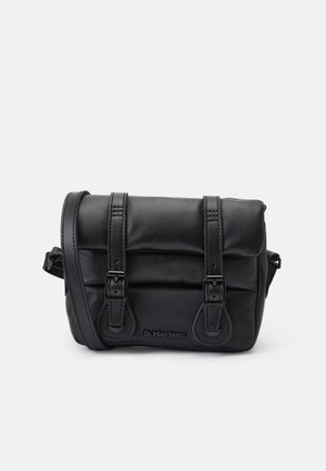 Sac à épaule en cuir noir avec deux sangles à boucle et un logo de marque embossé sur le devant sur un fond clair uni.