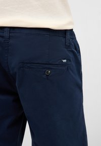 Gros plan sur un pantalon bleu marine avec une poche arrière boutonnée et des passants de ceinture, porté avec une chemise claire rentrée.