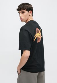 Fekete, rövid ujjú póló, melyen színes, retró stílusú grafikai mintázat található a hátsó oldalon, egyszínű háttér előtt kiállítva.