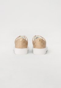 Bežīgas slip-on kurpes ar teksturētu auduma augšdaļu, baltām gumijas zolēm, logotipa akcentiem un vienkāršu dizainu. Attēlots aizmugures skats.