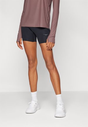 Kortermet, mauve treningsgenser over tettsittende svarte shorts; akzentuert med en logo på shortsen. Hvite joggesko og ribbede ankelstrømper synlige.