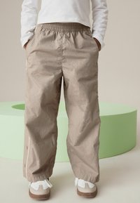 Pantalons légers couleur sable avec une taille élastique, des poches latérales et des fermetures à pression au niveau de l'ourlet. Portés avec des baskets blanches et grises.