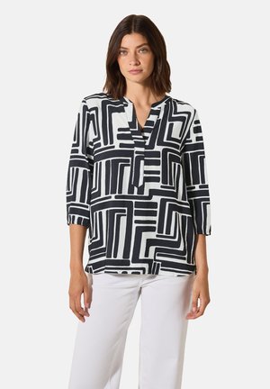 Blouse à motifs noirs et blancs avec un col en V, des manches trois-quarts et des motifs géométriques sur un tissu léger. Associée à un pantalon blanc.