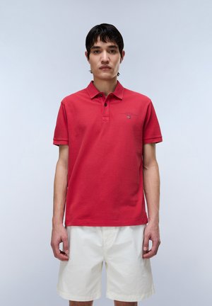 Jeune homme portant un polo rouge avec un petit logo et un short blanc jusqu'aux genoux, debout devant un fond gris clair uni.