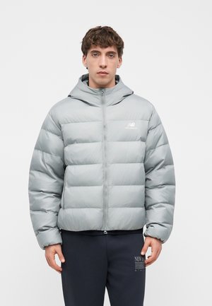 Junger Mann trägt eine hellgraue New Balance Daunenjacke mit Kapuze und steht vor einem schlichten weißen Hintergrund.