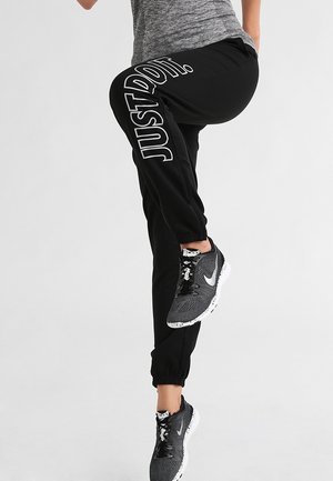 Personne portant un pantalon de survêtement noir avec l’inscription « JUST DO IT. » et des chaussures de sport grises, levant une jambe sur un fond uni.