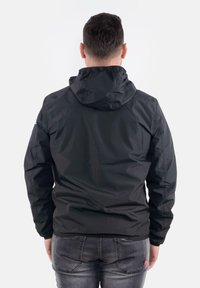 Giacca nera leggera con cappuccio, caratterizzata da una texture liscia e polsini elastici, indossata sopra jeans in denim grigi.