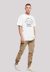 Hvit bomulls T-skjorte med grafisk trykk og løs passform, kombinert med beige cargo-bukser med flere lommer, og hvite sneakers.