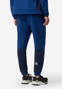 Niebieskie spodnie sportowe z fakturowanym ciemnoniebieskim panelem dolnym, elastycznymi mankietami i detalem z logo w okolicach kostki. Noszone z czarnymi butami sportowymi.