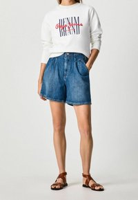 Felpa bianca con scritte rosse e blu, abbinata a pantaloncini di jeans blu con tasche laterali. Sandali marroni con cinturini completano il look.