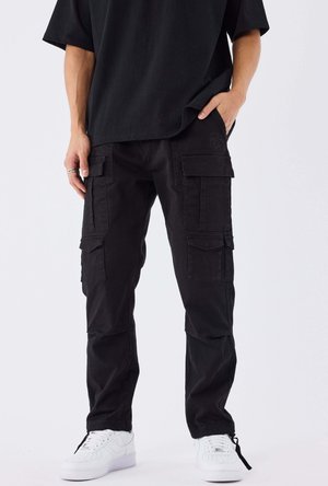 Persoon draagt zwarte cargobroek met meerdere zakken, een zwart shirt en witte sneakers, staat met één hand in de zak.