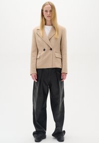 Beige dvouřadý kabát vyrobený z měkké tkaniny s černými knoflíky, spárovaný s širokými černými koženými kalhotami a černými botami.
