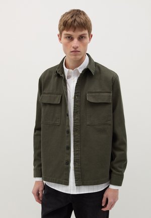 Chaqueta de camisa de algodón verde olivo con botones en la parte delantera, dos bolsillos en el pecho y un dobladillo recto, que se lleva sobre una camisa blanca con cuello.