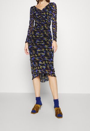 Femme portant une robe froncée à manches longues avec un imprimé abstrait noir, bleu et jaune, associée à des chaussettes bleues et des talons ouverts marron.