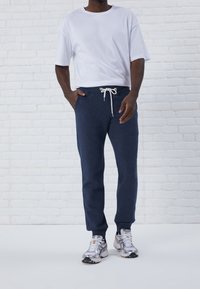 Mörkblå sweatpants med vit dragsko, matchade med en figurnära vit t-shirt och silverfärgade sportskor. Står mot en vit tegelvägg.