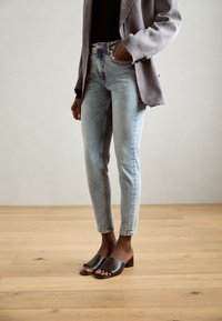 Jeans azzurri chiari con un design aderente, abbinati a un blazer grigio e mule scure con punta aperta su uno sfondo di pavimento in legno.