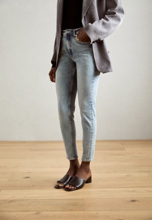Lyseblå jeans med en tettsittende design, kombinert med en grå blazer og mørkebrune åpne mules på en bakgrunn av tregulv.