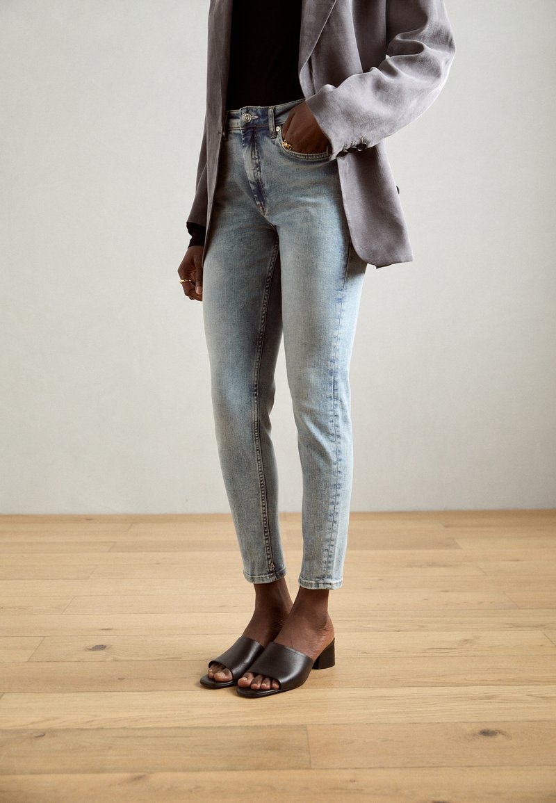 Jeans azzurri chiari con un design aderente, abbinati a un blazer grigio e mule scure con punta aperta su uno sfondo di pavimento in legno.