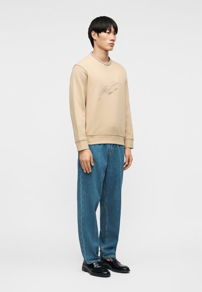 Νεαρός άνδρας που φοράει μπεζ φούτερ με λογότυπο, μπλε τζιν και μαύρα παπούτσια, στέκεται μπροστά σε απλό ανοιχτό γκρι φόντο.