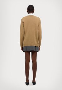 Pull beige oversize avec un col rond, des poignets et un ourlet côtelés. Porté par-dessus une jupe plissée grise, vu de dos.