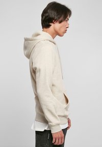 Beige hettegenser med kenguro-lomme, ribbede mansjetter og kant. Myk tekstur, uformell design. Båret over en hvit skjorte.