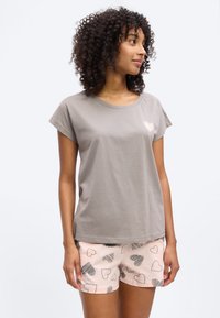 Γυναίκα με σγουρά μαλλιά που φοράει γκρι κοντομάνικο πουκάμισο με μια μικρή καρδιά στο στήθος και ροζ σορτς με μοτίβα καρδιών σε μαύρο και άσπρο.