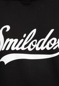 Sudadera negra con gran texto en cursiva blanca "Smilodox" en la parte frontal. El material es suave, con un diseño clásico de capucha.