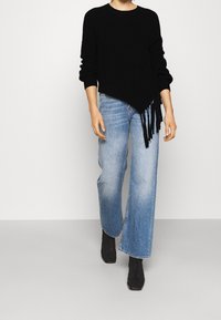 Sort strikket sweater med diagonal kant og frynser på den ene side, parret med lyseblå vidde jeans og sorte ankelstøvler.