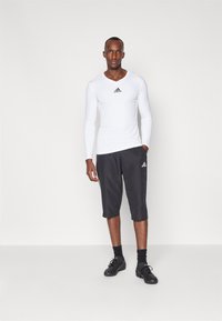 Weißes Langarmhemd mit schwarzem Logo, kombiniert mit schwarzen knielangen Shorts mit weißen Akzenten. Getragen mit schwarzen Sportschuhen und Socken.
