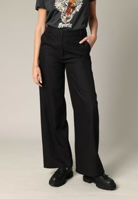 Pantalons larges noirs à taille haute, en tissu lisse. Le modèle porte un t-shirt graphique et des chaussures noires chunky.